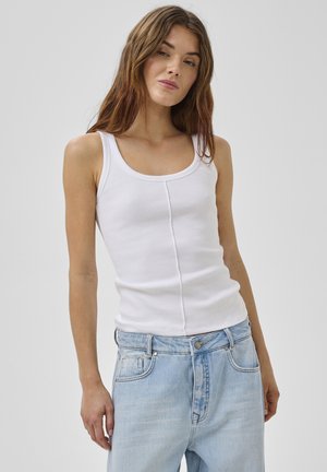 Jonge vrouw met lang bruin haar, gekleed in een witte mouwloze geribbelde top en lichtblauwe high-waisted spijkerbroek, tegen een effen achtergrond.