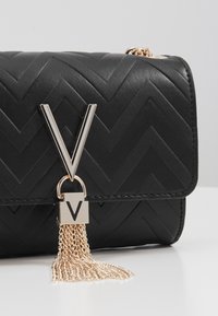 Czarna skórzana torebka z wzorem w jodełkę, srebrnymi elementami, dużą literą „V” i zwisem z łańcuszka. Kompaktowy, prostokątny kształt.