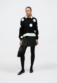 Camisola preta com bolinhas brancas, com uma camisa branca de gola por baixo, combinada com uma saia preta plissada e meias pretas. Acessórios incluem uma mala preta.