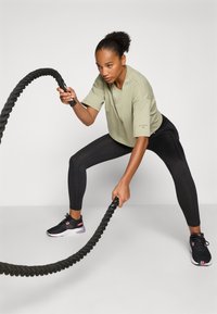 Grünes Baumwoll-Crop-Top, schwarze Leggings und schwarze Sportschuhe mit pinken Akzenten. Hält ein verdrehtes schwarzes Battle Rope in breiter Stellung.