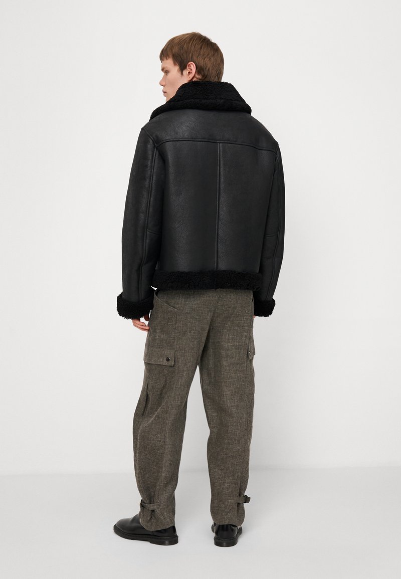 Chaqueta de cuero negra con un cuello y puños de piel de oveja negra, combinada con pantalones grises de corte holgado y textura, y botas negras.