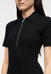 Črna rebrasta obleka s kratkimi rokavi, z ostrim ovratnikom in srebrnim polovičnim zadrgo. Teksturirana tkanina s prilegajočo se obliko in gladkim zaključkom.