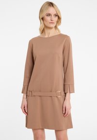 Frau in einem knielangen, beige-farbenen Kleid mit langen Ärmeln und zwei rechteckigen Schnallen nahe dem Saum vor einem weißen Hintergrund.