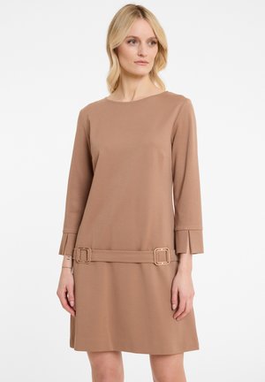 Femme portant une robe couleur camel, longueur genoux, avec des manches longues et deux boucles rectangulaires près de l'ourlet, sur fond blanc.