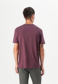 Bordeauxfarbene Kurzarm-T-Shirt mit Rundhalsausschnitt, aus einem weichen, leichten Stoff hergestellt und mit einem lockeren Sitz. Sichtbares Tattoo am linken Arm.