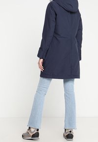 Marinblå parka med huva, med rak skärning, sidofickor och justerbara ärmmuddar. I kombination med ljusblå utsvängda jeans och grå skor.