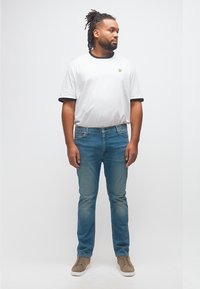 Homme avec dreadlocks portant un t-shirt blanc avec des bordures noires, un jean bleu et des baskets beiges, debout et regardant sur le côté sur un fond uni.