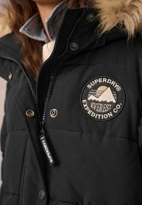 Gros plan d'une personne portant une veste matelassée noire Superdry avec capuche bordée de fourrure et écusson brodé Everest Expedition Co.