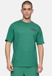 Groene t-shirt met korte mouwen van katoen. Heeft paarse geborduurde "R.S.V.P." op de linkerborst. Rechte snit met een ronde hals.