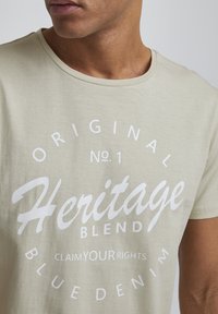 Bež T-shirt od pamuka s bijelim natpisom. Dizajn s okruglim izrezom i kružnom tipografijom: "Heritage Blend," "Claim YOUR Rights," i "Blue Denim."