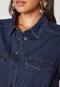 Camicia di denim blu scuro con colletto classico, bottoni a pressione e due tasche sul petto; presenta cuciture a contrasto e una texture liscia.