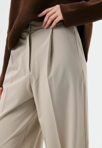 Beige, op maat gemaakte broek met hoge taille, voorzien van plooien aan de voorkant en zijzakken, gemaakt van gladde stof. Bruine trui gedeeltelijk zichtbaar.