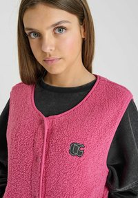 Roze fleecevest met een gestructureerd oppervlak, ronde halslijn en knopen aan de voorkant. Beschikt over een logopatch op de borst.