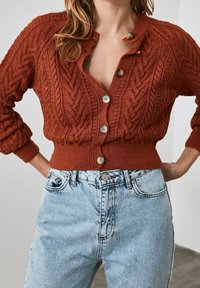 Kabelstrick-Cardigan in rostrot mit tiefem V-Ausschnitt, gerippten Bündchen und Knopfleiste mit fünf Knöpfen, kombiniert mit hellblauen, hoch taillierten Jeans.