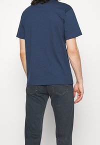 Krátké tričko s krátkým rukávem v námořnické modři s kulatým výstřihem, zobrazené zezadu. Materiál vypadá měkký a hladký, s pohodlným střihem.