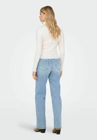 Kobieta z długimi blond włosami, ubrana w białą bluzkę z długim rękawem, jasnoniebieskie proste dżinsy i brązowe botki na obcasie, stojąca tyłem.