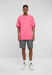 T-shirt rose surdimensionné avec un col rond, associé à un short cargo gris. Le mannequin porte des baskets blanches avec des accents noirs et des chaussettes blanches.