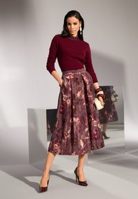 Frau steht mit der Hand in der Tasche und trägt ein burgunderfarbenes Langarmoberteil, einen floralen, plissierten Midirock, burgunderfarbene Schuhe und hält eine weiße Clutch sowie Armbänder.