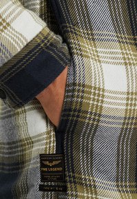 Camisa a cuadros en verde, azul marino y blanco con una textura cepillada. Presenta una etiqueta tejida, detalles de botones y materiales contrastantes en el cuello y los puños.