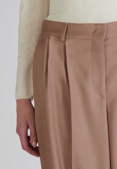 Brune op maat gemaakte broeken met plooien, gemaakt van een gladde stof en een platte tailleband. Een hand raakt de zijnaad aan. Een lichtgekleurde top is gedeeltelijk zichtbaar.