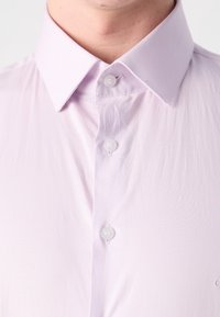 Gros plan d'une personne portant une chemise boutonnée violet clair avec un col pointu, mettant l'accent sur le col et les boutons supérieurs.