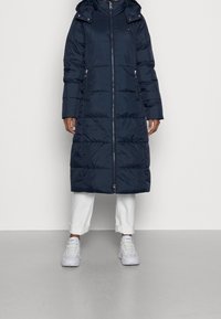 Tmavě modrý péřový kabát s kapucí, celým zipem a bočními kapsami. Má prošívaný vzor a střední délku, doplněný bílými kalhotami a teniskami.