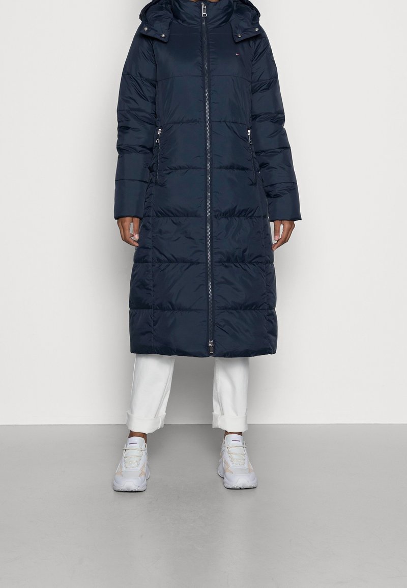 Tmavě modrý péřový kabát s kapucí, celým zipem a bočními kapsami. Má prošívaný vzor a střední délku, doplněný bílými kalhotami a teniskami.