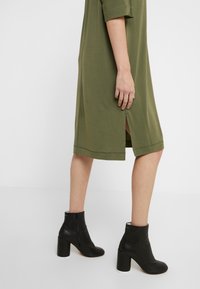 Robe vert olive avec des manches courtes et des fentes sur les côtés, associée à des bottines en cuir noire à talon épais et fermeture éclair sur le côté.