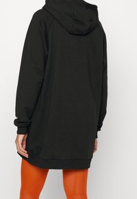 Robe à capuche noire avec une capuche spacieuse, des manches longues et des poignets côtelés. Offre une coupe décontractée et une texture douce. Fentes latérales visibles au ourlet.