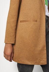 Brun kappa i ullblandning med strukturerad design, med en enkel bröstficka och slät yta. Bärs över mörka jeans.