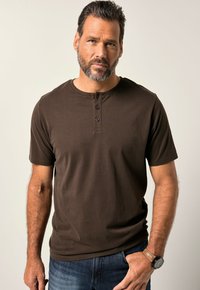 T-shirt Henley marrone a maniche corte con scollatura a girocollo e patta con tre bottoni. Tessuto morbido, abbinato a jeans in denim blu e un orologio.