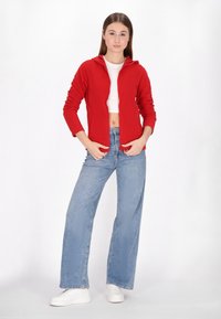 Czerwona bluza z kapturem na zamek, noszona na białym topie crop, w połączeniu z jasnoniebieskimi spodniami typu wide-leg i białymi trampkami. Gładka tekstura materiału.