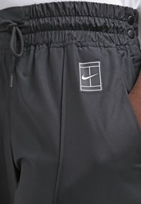 Black athletic pants featuring an elastic waistband with adjustable drawstring, side pockets, and a white Nike logo on the thigh area.