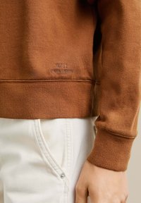 Sudadera de algodón marrón con puños y cinturilla acanalados, y logo bordado. Combinada con jeans blancos que presentan un detalle de botones.