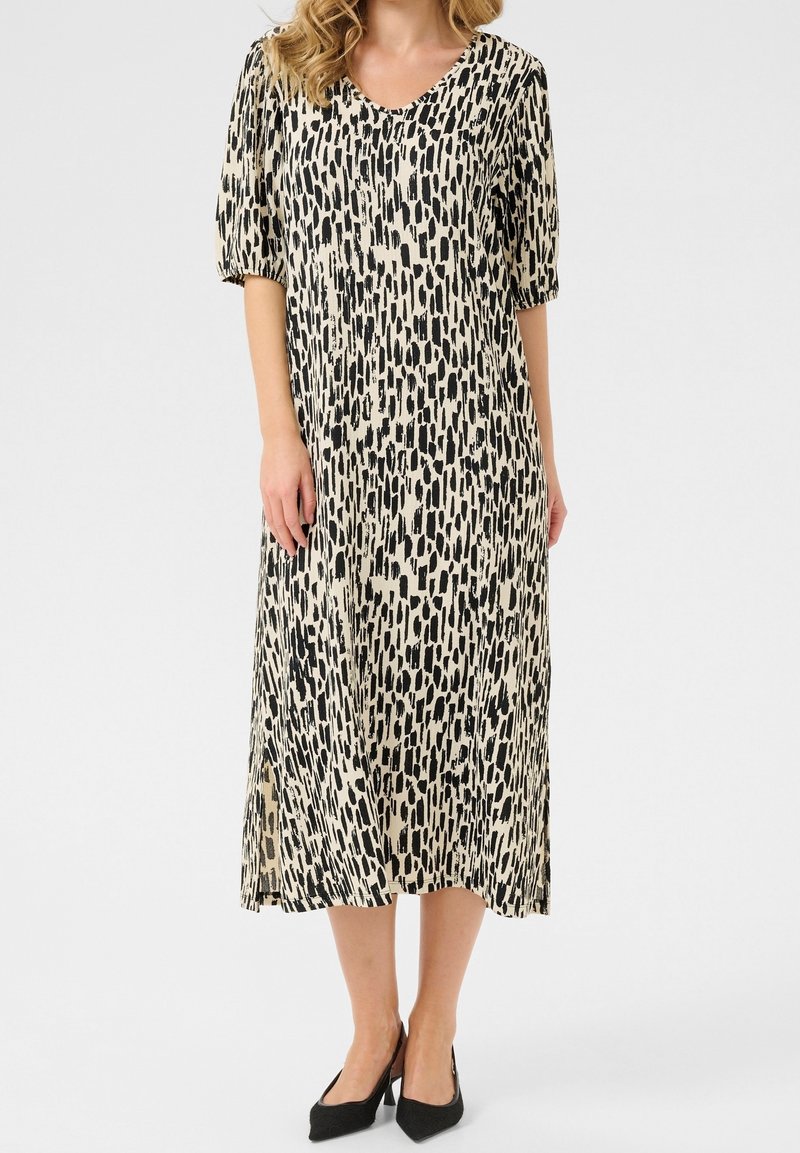 Femme portant une robe midi beige avec un motif abstrait de coups de pinceau noirs et des manches bouffantes jusqu'aux coudes, associée à des talons pointus noirs.