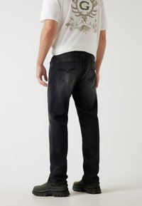 Schwarze Denim-Jeans mit geradem Schnitt, sichtbaren Gesäßtaschen und kontrastierenden Nähten. Kombiniert mit einem weißen Grafik-T-Shirt und dunklen Stiefeln.