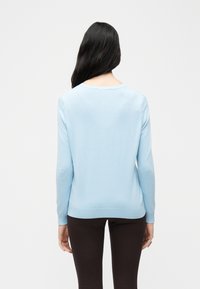 Γυναίκα με μακριά σκουρόχρωμα μαλλιά, ντυμένη με ανοιχτό μπλε πουλόβερ με μακριά μανίκια και σκούρο παντελόνι, που στέκεται με την πλάτη προς τα εμάς μπροστά σε απλό φόντο.