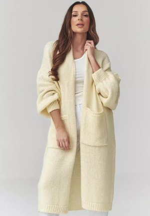 Kobieta ubrana w długi, kremowy, gruby sweter z kieszeniami z przodu, na białej bluzce i spodniach, stojąca na jednolitym tle.