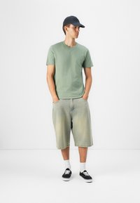 Tricou verde din bumbac, şorturi din denim deschis, capac negru, ciorapi albi, adidaşi negri, ținută casual cu o croială relaxată și texturi uzate.