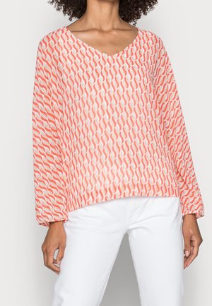 Femme portant un chemisier à manches longues corail et blanc à motif, col en V, avec un pantalon blanc ample sur un fond uni.