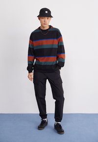 Gestreepte sweatshirt met zwart, rood, groen en blauwe kleuren. In combinatie met zwarte cargo broeken en een zwarte bucket hat. Casual schoeisel inbegrepen.