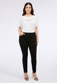 Camiseta blanca de manga corta, con diseño de texto en el frente, combinada con jeans negros ajustados y tacones negros con tiras. Tela suave, estilo casual.