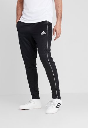 Mężczyzna ubrany w czarne spodnie dresowe Adidas z białym paskiem i białe wysokie trampki Adidas, stojący z rękami w kieszeniach.