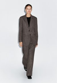 Donna che cammina in avanti indossando un completo di lana marrone scuro con scarpe nere e un top nero, su uno sfondo bianco uniforme.