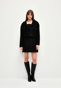 Schwarze Tweed-Shortjacke mit zwei vorderen Taschen, kombiniert mit einem schwarzen Kleid. Das Model trägt kniehohe schwarze Stiefel. Glatte Textur, strukturiertes Design.