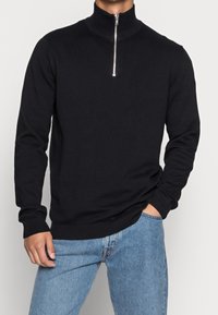 Pull en tricot noir avec un col montant et une fermeture éclair argentée. Doté de poignets et d'un ourlet côtelés, porté avec un jean bleu clair. Design simple.