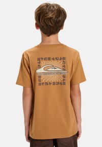 Camiseta de algodón marrón con mangas cortas. El diseño en la parte posterior presenta un logo y patrones abstractos en gris y crema sobre un fondo de rayos de sol.