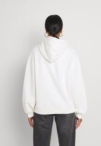 Mujer con el cabello oscuro recogido, usando pendientes de aro dorados, un sudadera blanca holgada y pantalones gris oscuro, de espaldas ante un fondo sencillo.
