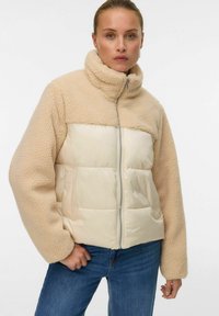 Sieviete, kas valkā bēša krāsas sherpa flīsa un polsterētu jaku ar augstu apkakli, kombinācijā ar ziliem džinsiem, stāvot pret baltu fonu.