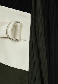Geribbelde stof in zwart, olijfgroen en crème, met een brede crèmekleurige riem met een ring van zilver metaal, wat een kleurgeblokkeerd ontwerp creëert.